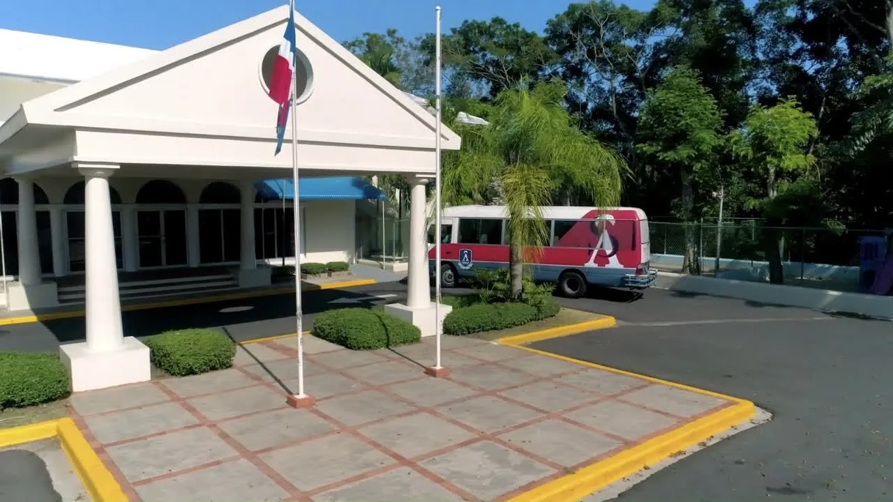 Entrada del American School of Santo Domingo con la bandera dominicana y un autobús del colegio, en Arroyo Hondo