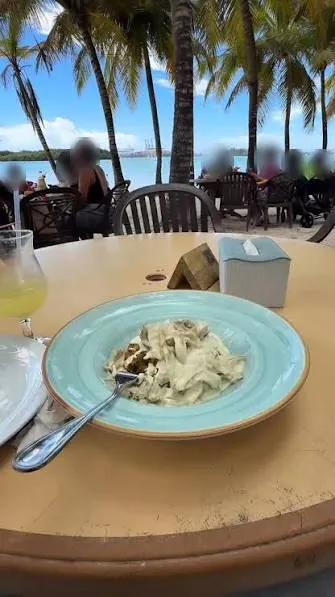 Plato de pasta en una mesa de Bocana Beach, con palmeras, el mar y las grúas de Caucedo al fondo