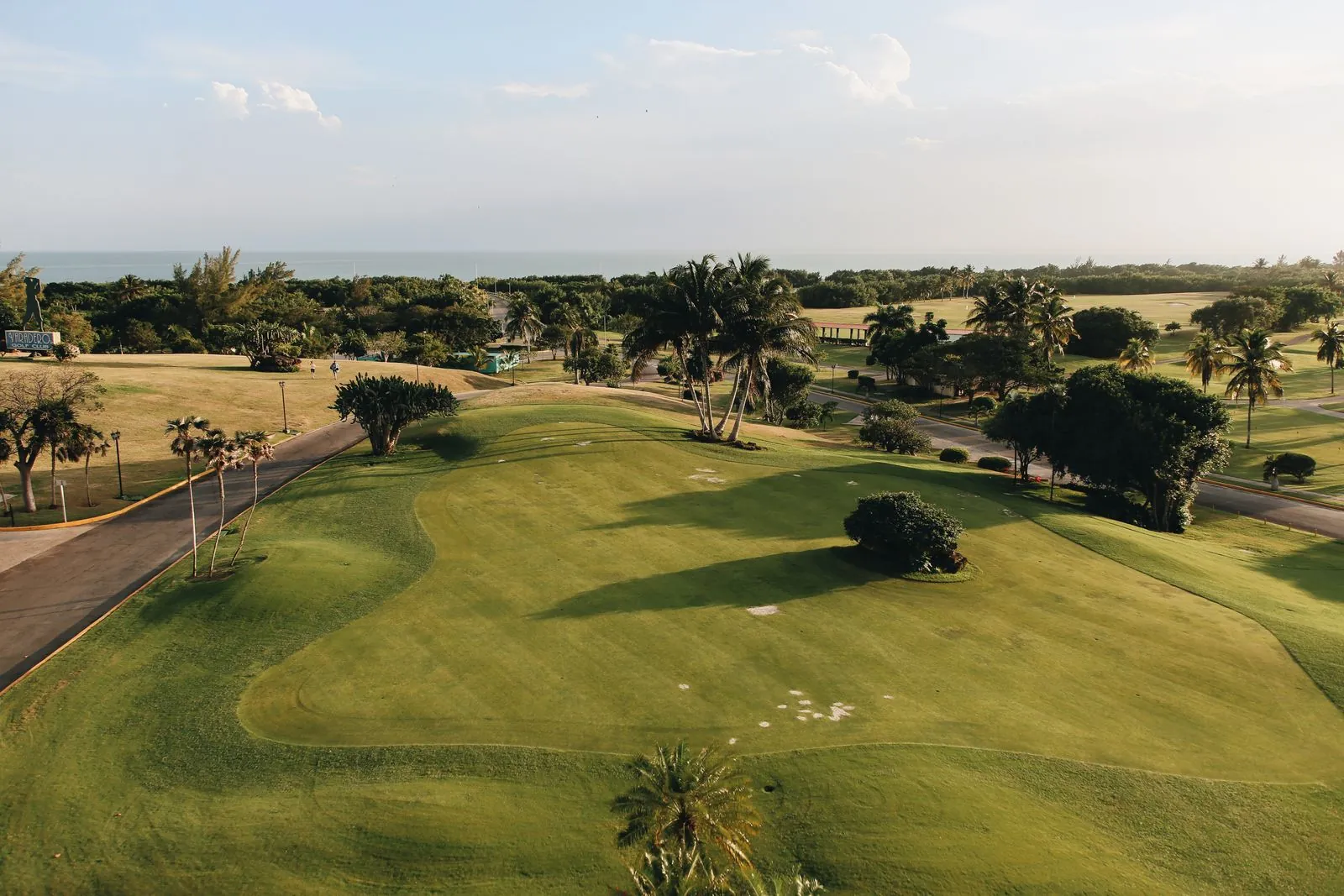 Vista aérea de un campo de golf entre palmeras junto al mar, con sus calles de césped y trampas de arena