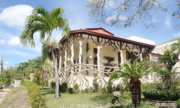 Casa típica de Gazcue, Santo Domingo, de una planta con techo rojo y una galería decorada con columnas en forma de troncos, rodeada de palmas