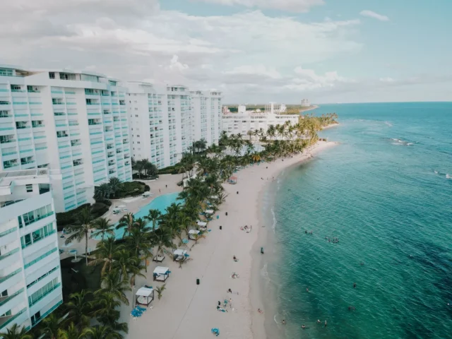 Vista aérea de la playa de Juan Dolio con torres de apartamentos blancas, piscinas y palmeras frente al mar Caribe | Las torres de apartamentos de Juan Dolio, con sus piscinas y palmeras, a pocos pasos de la playa. Así se ve hoy la franja frente al mar, donde los hoteles de antes dieron paso a la segunda residencia. | Abel Acosta / Pexels