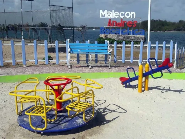 Letrero «Malecón Andrés Bocachica» con un banco y juegos infantiles sobre la arena, frente a la bahía