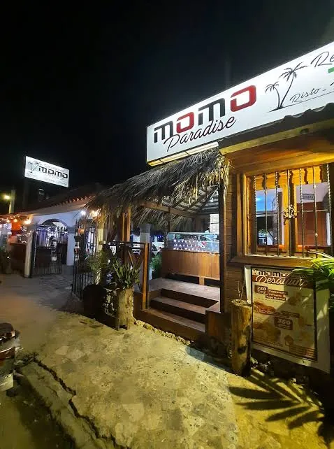 Fachada de Momo Paradise de noche, con su letrero iluminado y la entrada de madera bajo un techo de guano