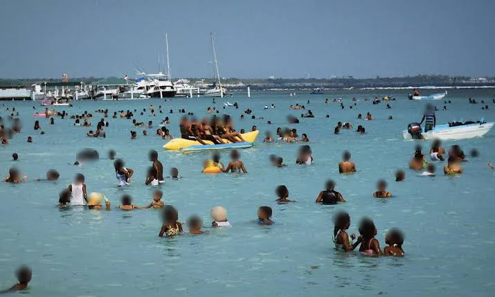 Cientos de bañistas en el agua poco profunda de la playa Boca Chica, con una banana inflable y yates al fondo