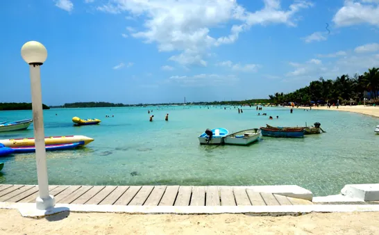 Barcas, lanchas y una banana inflable junto a un muelle de Boca Chica, con bañistas al fondo en la laguna