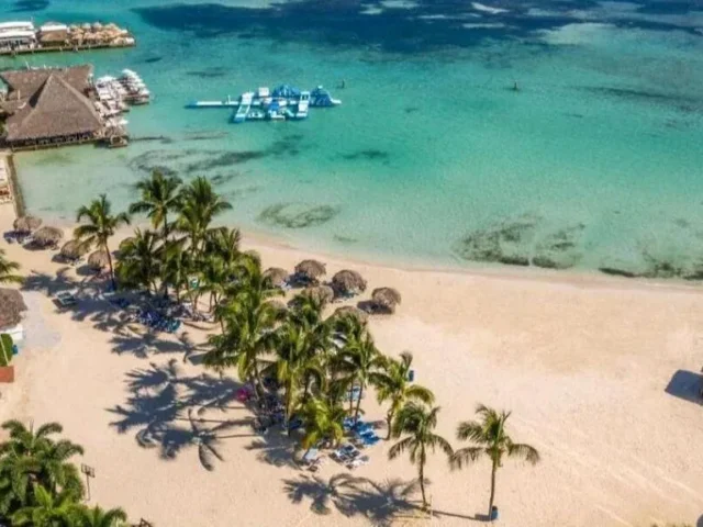 Vista aérea de la arena de Boca Chica con palmeras, sombrillas de guano, muelles de restaurantes y un parque inflable en el agua | Desde el aire se ve cómo las palmeras, las sombrillas y los muelles de los restaurantes rodean la laguna. Las manchas oscuras en el agua son parches de coral y algas.