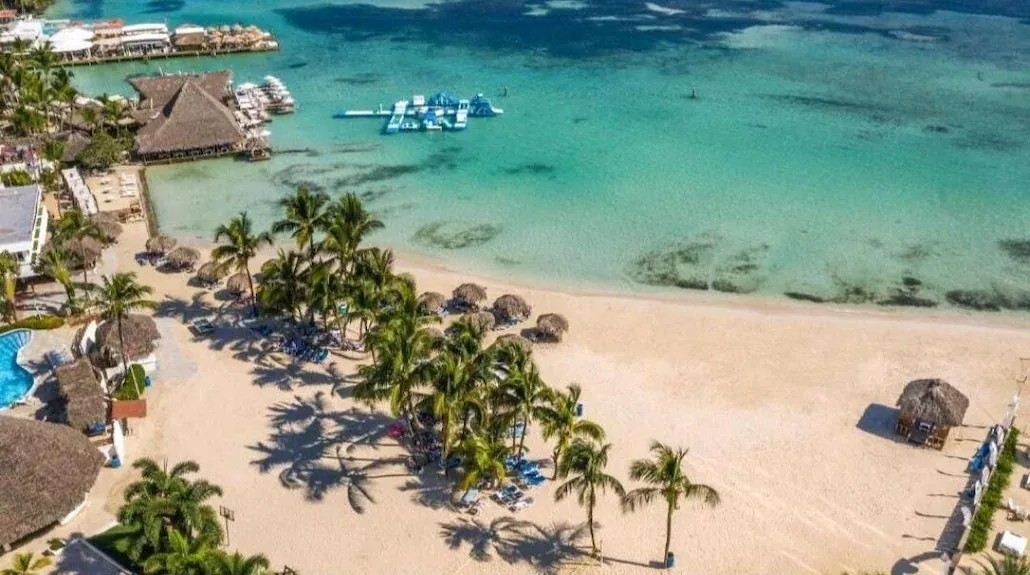 Vista aérea de la arena de Boca Chica con palmeras, sombrillas de guano, muelles de restaurantes y un parque inflable en el agua