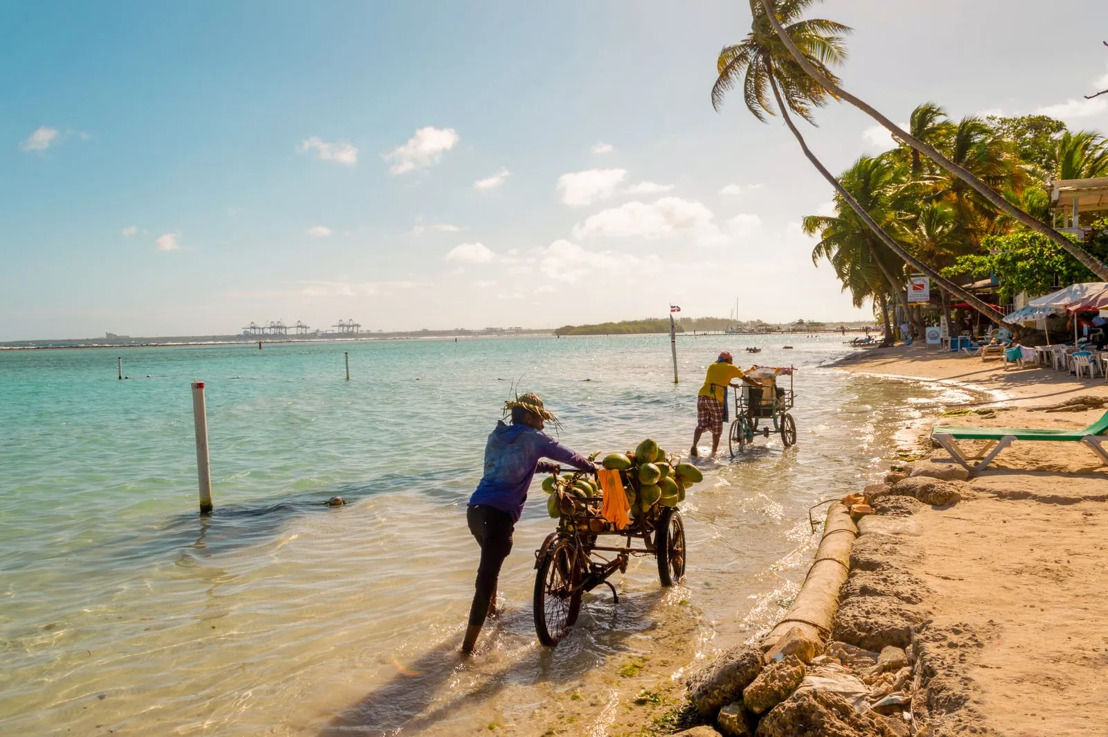 Dos vendedores empujan bicicletas cargadas por la orilla de una playa de aguas tranquilas en la República Dominicana